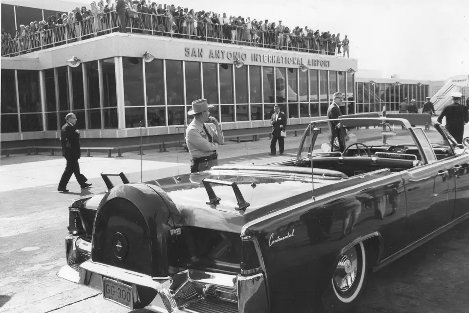 Black and white photo of President John F. Kennedy's car at San Antonio International Airport, surrounded by a crowd on November 21, 1963.